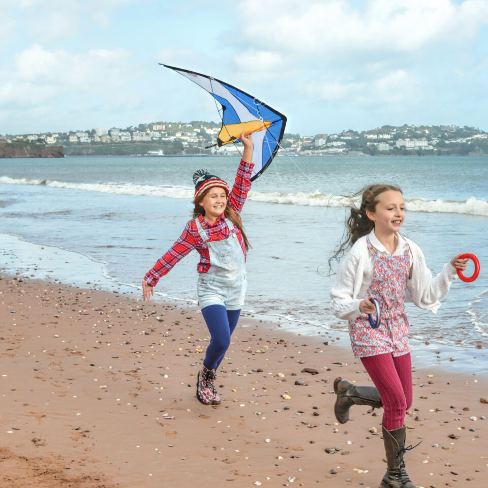 Two young girls running along a sandy beach with a blue and white kite.